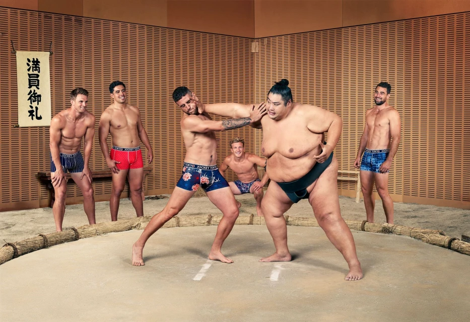 Group of muscular men posing in underwear with a sumo wrestler inside a sumo ring during a sporting event in an indoor environment.