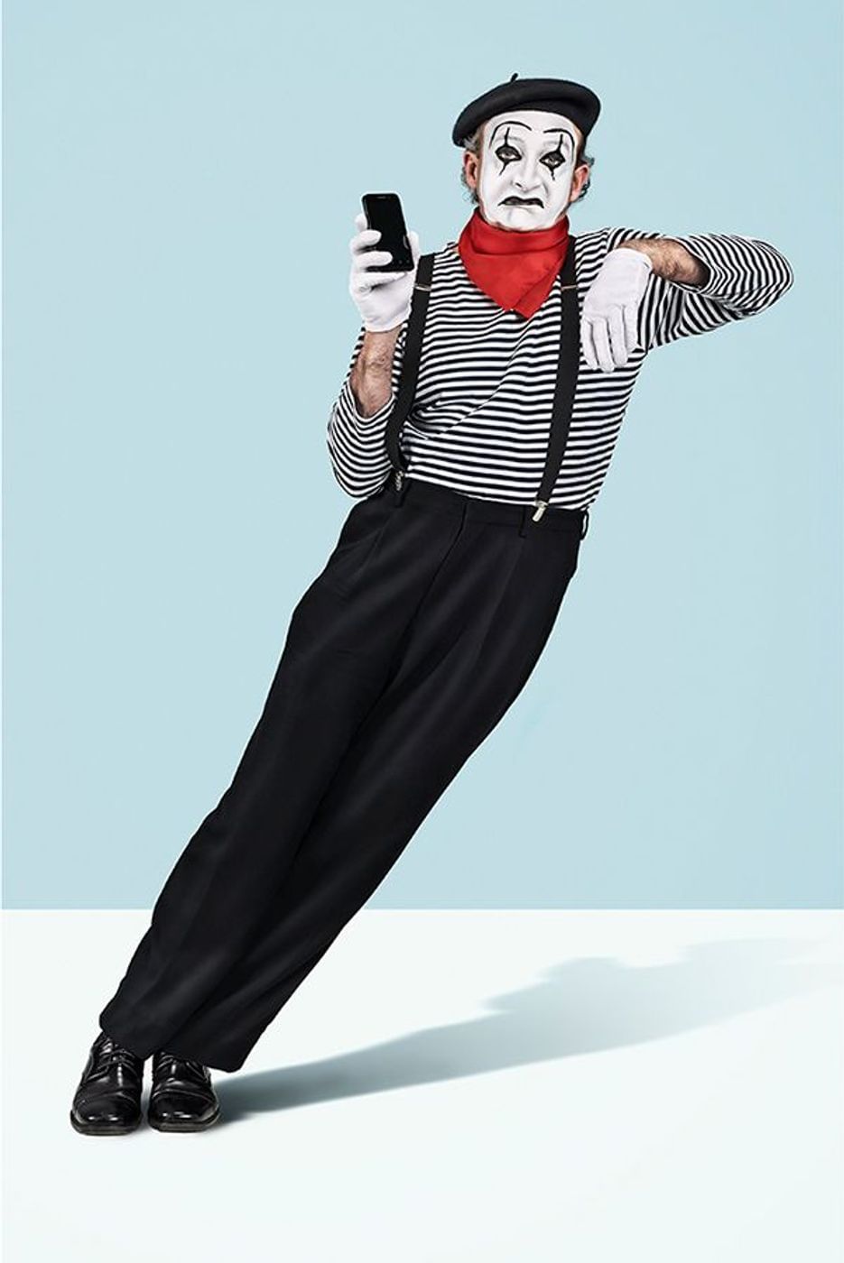 Full shot of a mime artist holding a smartphone, wearing a beret, striped shirt, black pants, gloves, and mime makeup in a studio.