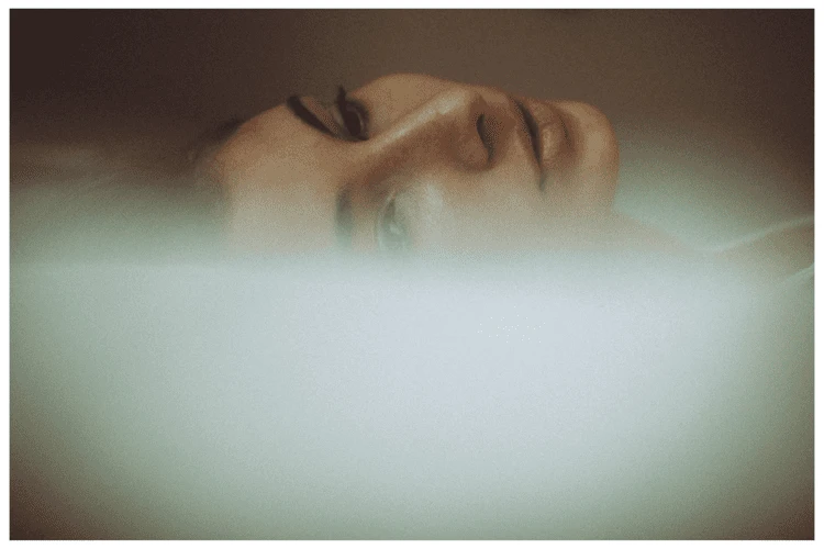 Close-up of an adult female face with a serene expression, partially submerged in what appears to be a milky substance or thick fog.