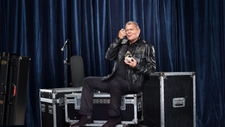 A mature man in a black leather jacket sitting on flight cases, eating dessert against blue velvet curtains.