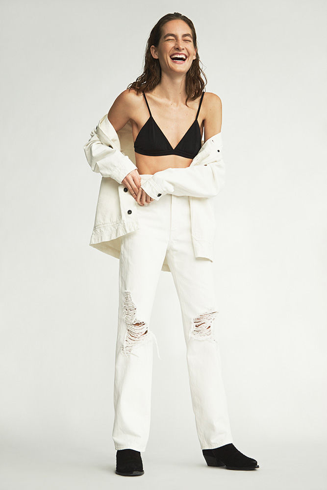 A full-length shot of a smiling, fair-skinned woman wearing a black bralette, white ripped jeans, and an unbuttoned white denim jacket.