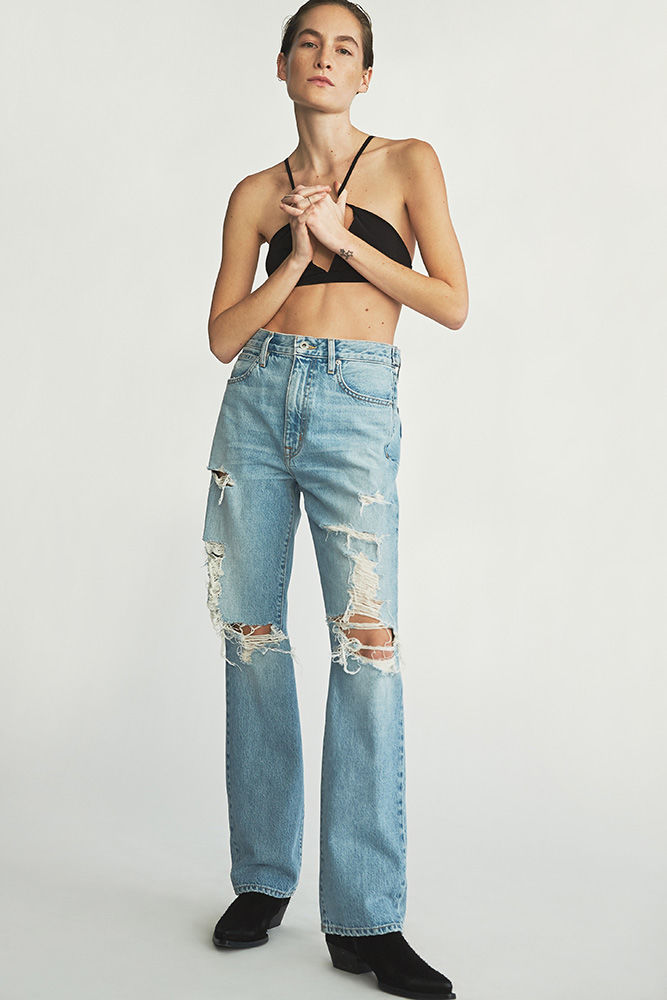 Full-length shot of a woman in a black halter top and light-wash, distressed denim jeans standing in front of a white backdrop.