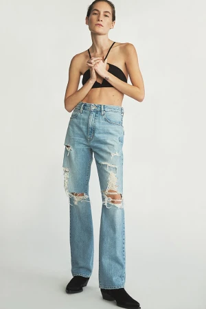 Full-length shot of a woman in a black halter top and light-wash, distressed denim jeans standing in front of a white backdrop.