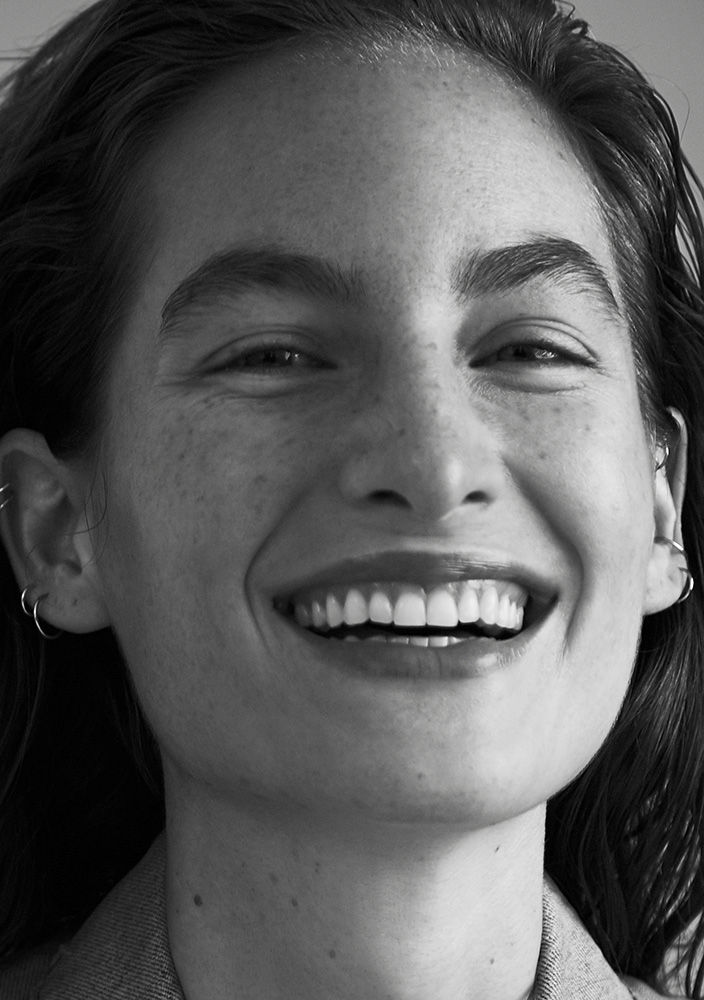 Close-up portrait of a smiling adult woman with freckles, wearing hoop earrings and a grey top, in a monochrome photograph.