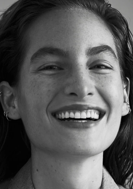 Close-up portrait of a smiling adult woman with freckles, wearing hoop earrings and a grey top, in a monochrome photograph.
