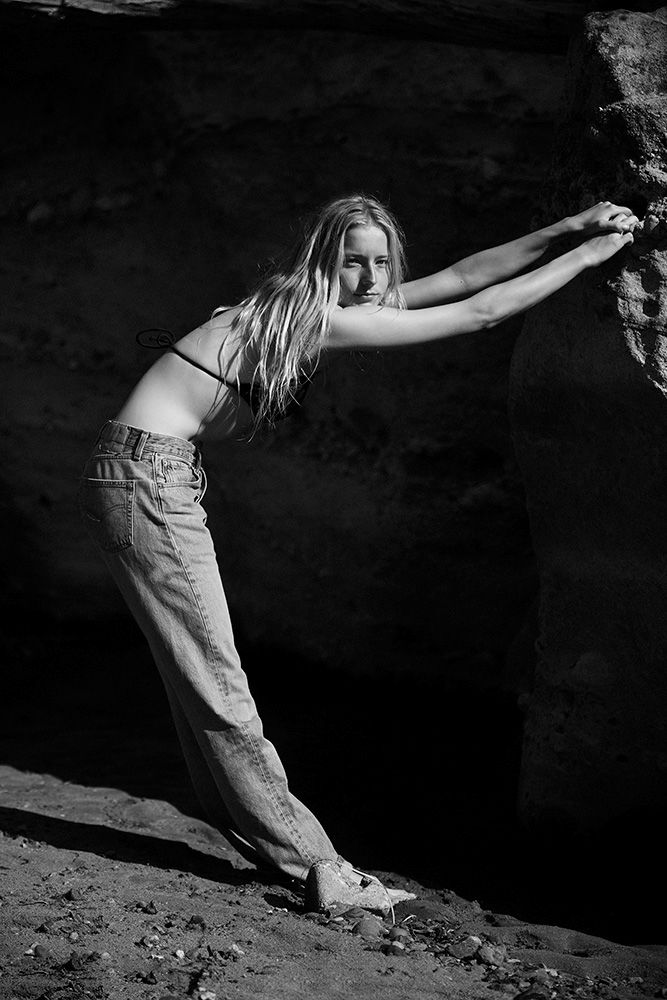 Monochrome portrait of a young woman leaning against a rocky outcrop wearing denim jeans and a bikini top