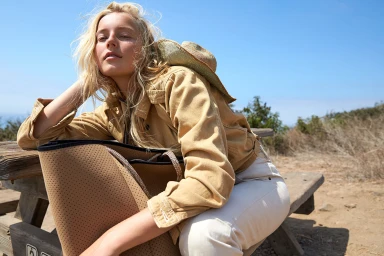 Blonde woman wearing a tan jacket and hat resting on a wooden picnic bench outdoors under a clear blue sky