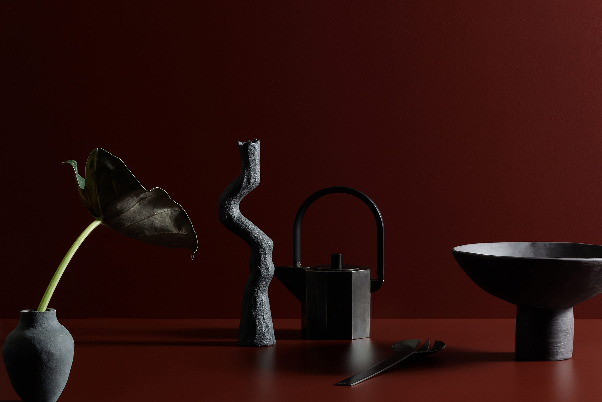 Still life featuring a dark green leaf in a gray vase, a gray sculpture, a dark tea pot, and a bowl against a dark red backdrop.