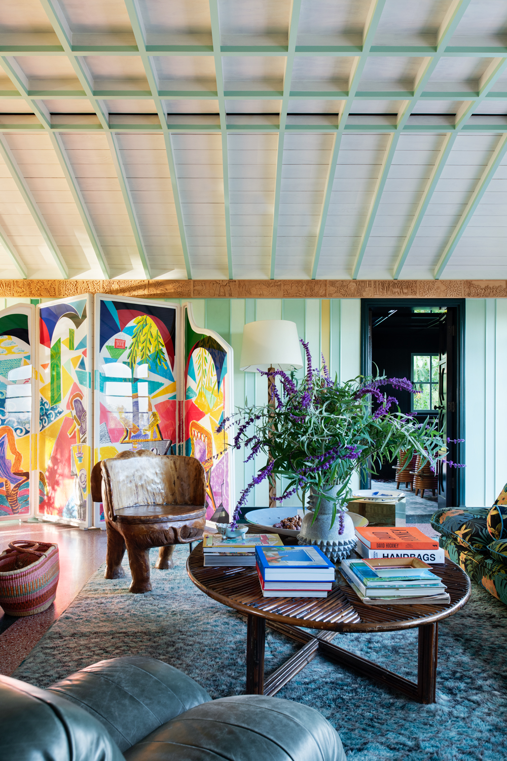 Interior view of a living room featuring colorful artwork, a unique wood chair, a woven table topped with books and flowers, and modern furnishings.