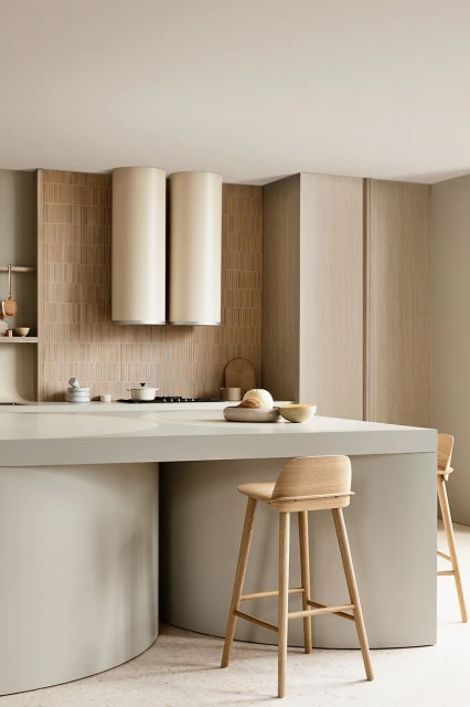 Beige kitchen interior with a curved island, wooden stools, modern cylindrical range hood, and neutral tile backsplash, illuminated by indoor lighting.