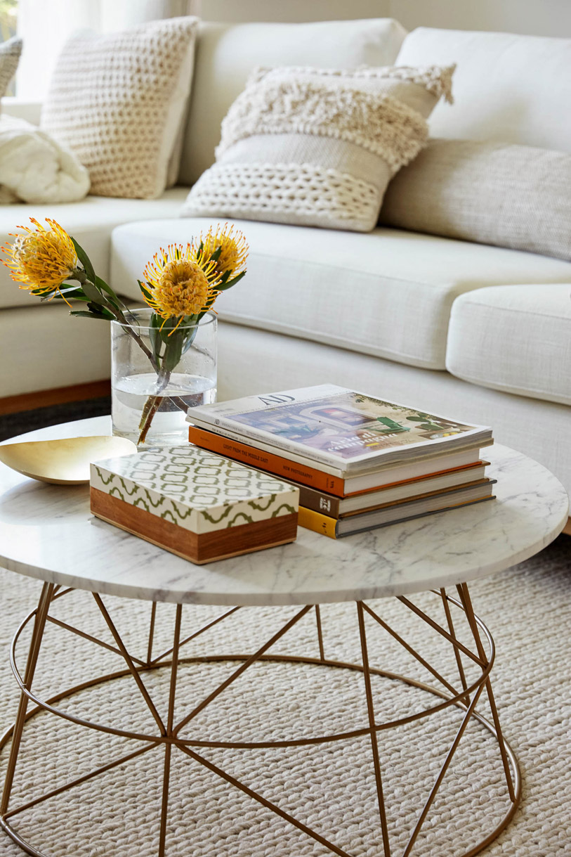 Living room interior with a white sofa, marble-top coffee table, floral arrangement, decorative books, and stylish pillows.