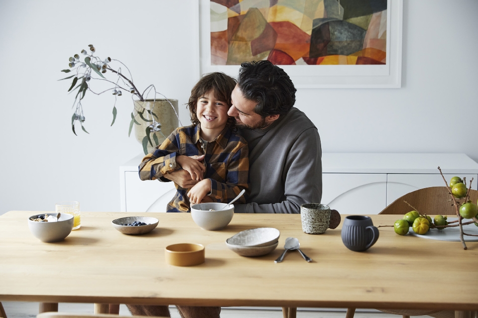 Smiling child and adult male embrace at a wooden table with bowls of fruit, juice, mugs, and ceramic tableware in a bright home interior.