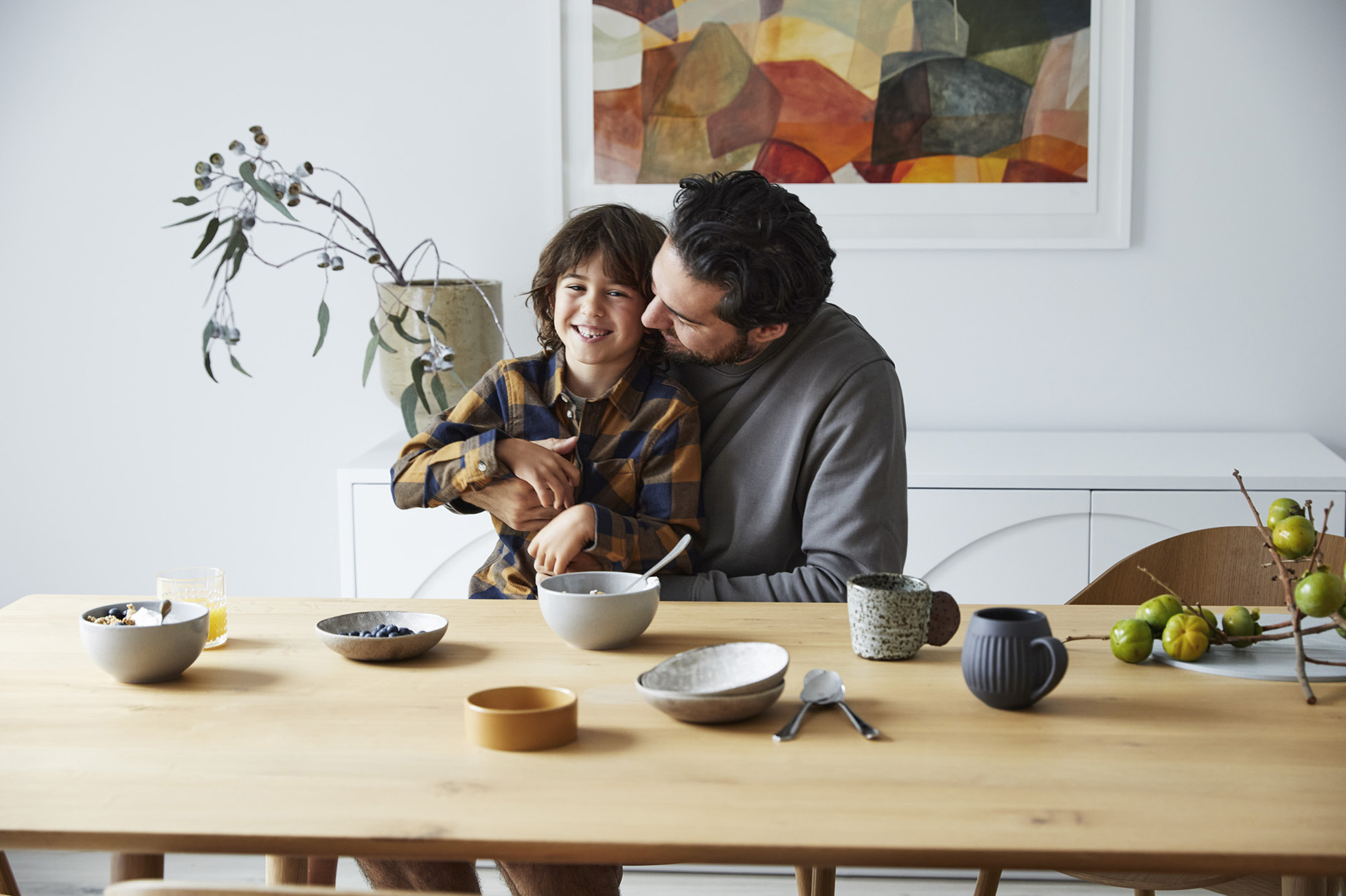 Smiling boy in a blue and brown plaid shirt embraces his father at a wooden dining table with fruit, cereal, and juice in an indoor setting.