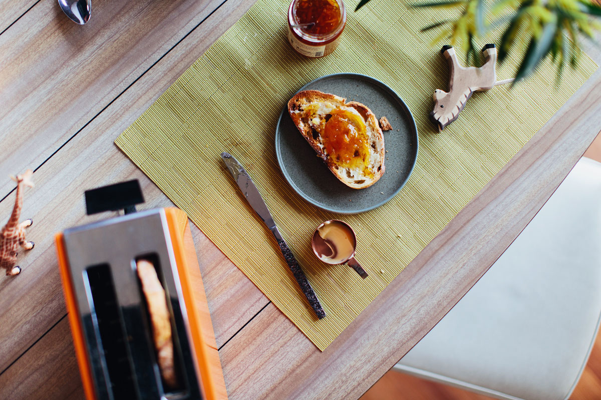 Overhead shot of a toast with marmalade, an orange toaster, utensils, and decorative animal figures on a wooden table with a green mat.