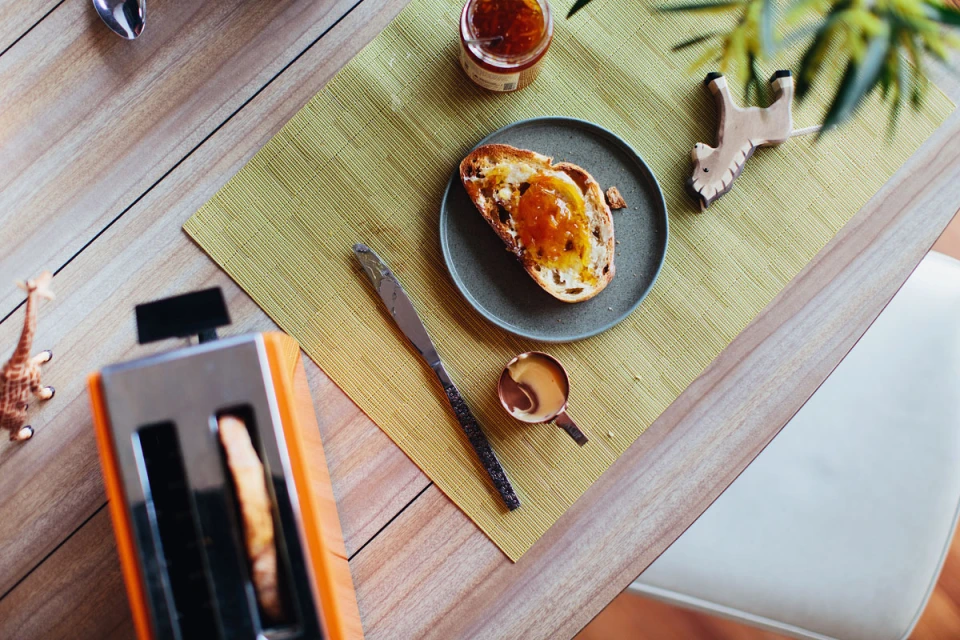 Overhead shot of a toast with marmalade, an orange toaster, utensils, and decorative animal figures on a wooden table with a green mat.