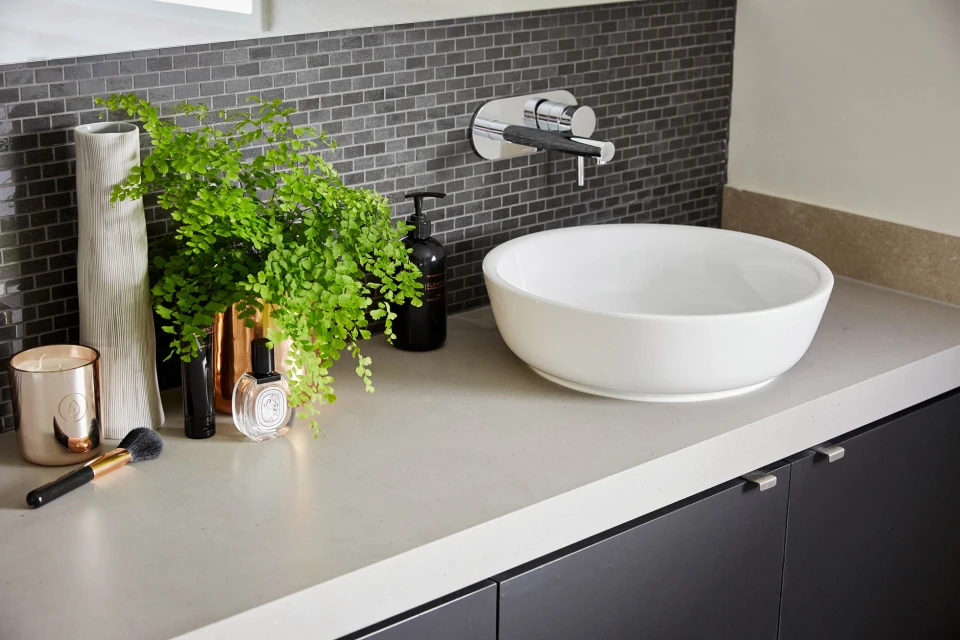 Modern bathroom interior featuring a white bowl sink, copper accents, leafy plant, and a dark grey tiled backsplash above the countertop.