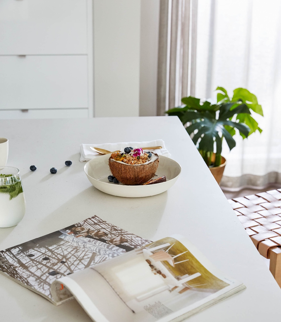 Overhead shot of a coconut bowl filled with granola, fruit, and edible flowers on a white table with a magazine and green beverage.