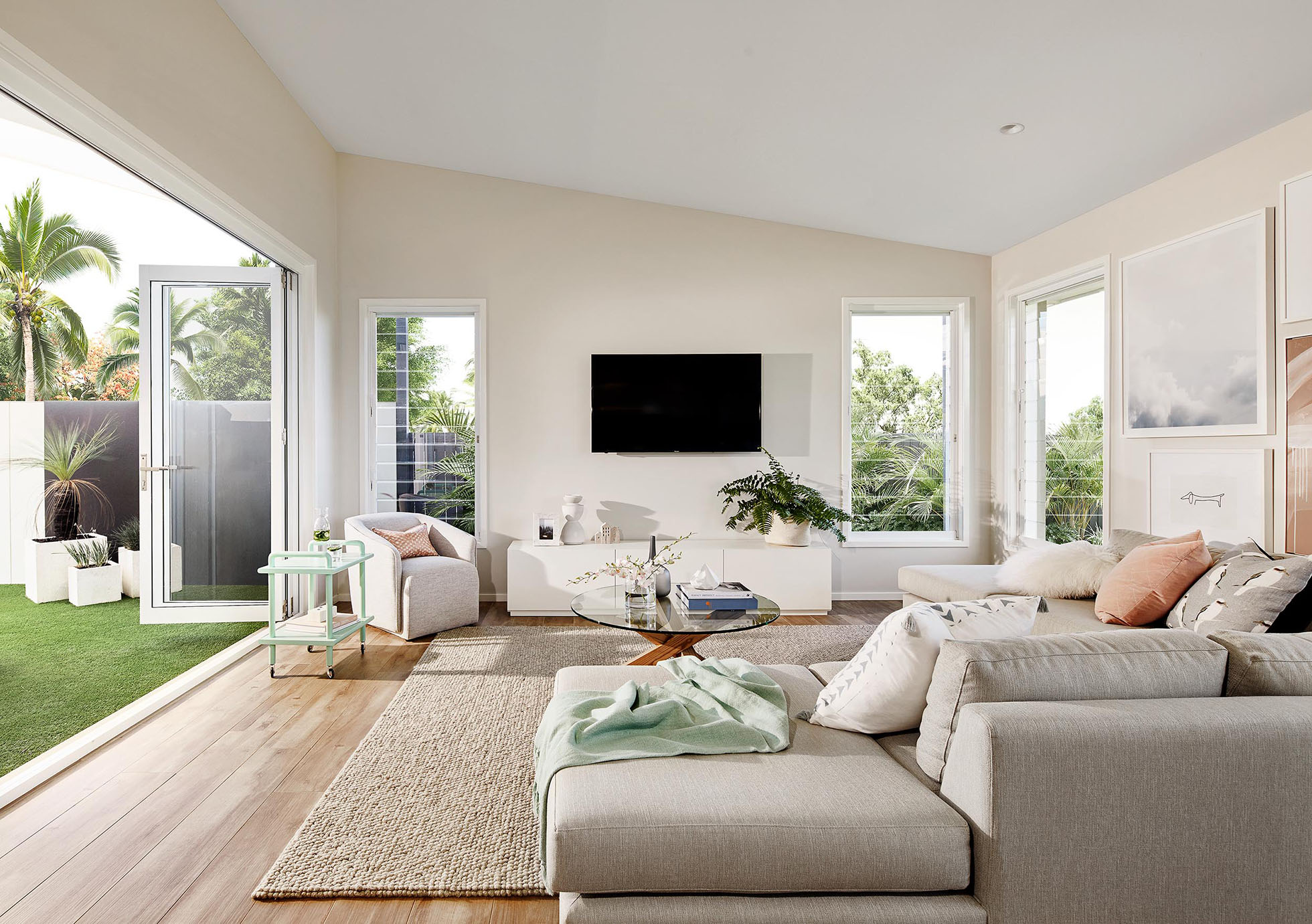 Bright living room interior with grey sectional sofa, round glass coffee table, and neutral rug. View of backyard with green lawn.