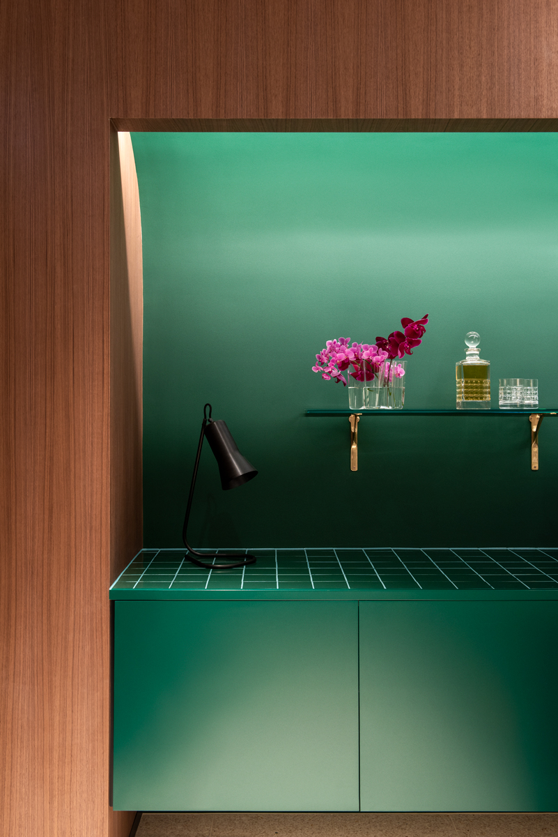 Stylish green and wood interior design featuring a tiled surface, table lamp, flowers, and glass decanter against a green backdrop.