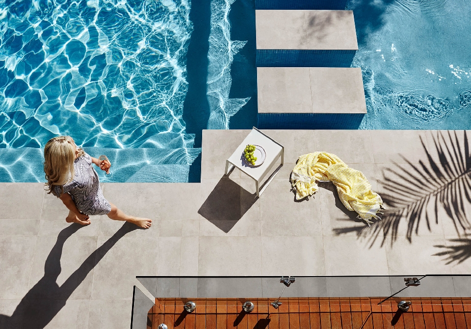 Overhead shot of a blonde woman walking barefoot by a pool with turquoise water, alongside a white table and yellow towel, outdoors.