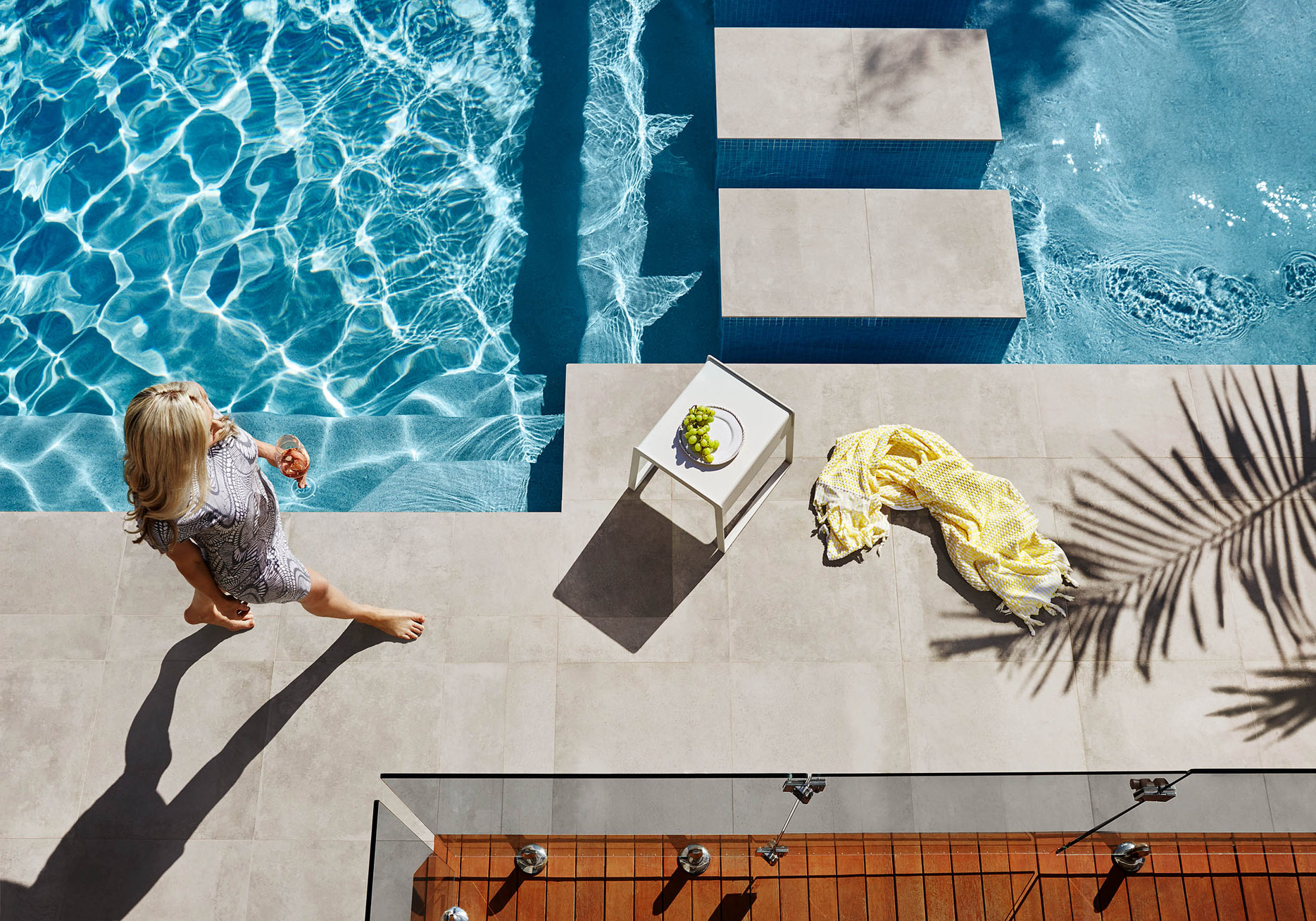 Overhead shot captures a blonde woman in a patterned dress next to a pool, with a table, grapes, towel, and palm frond shadow.