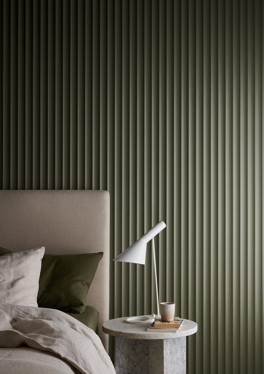 Interior shot of a bedroom with a bed featuring neutral-colored bedding and pillows, set against a vertically ridged green wall.