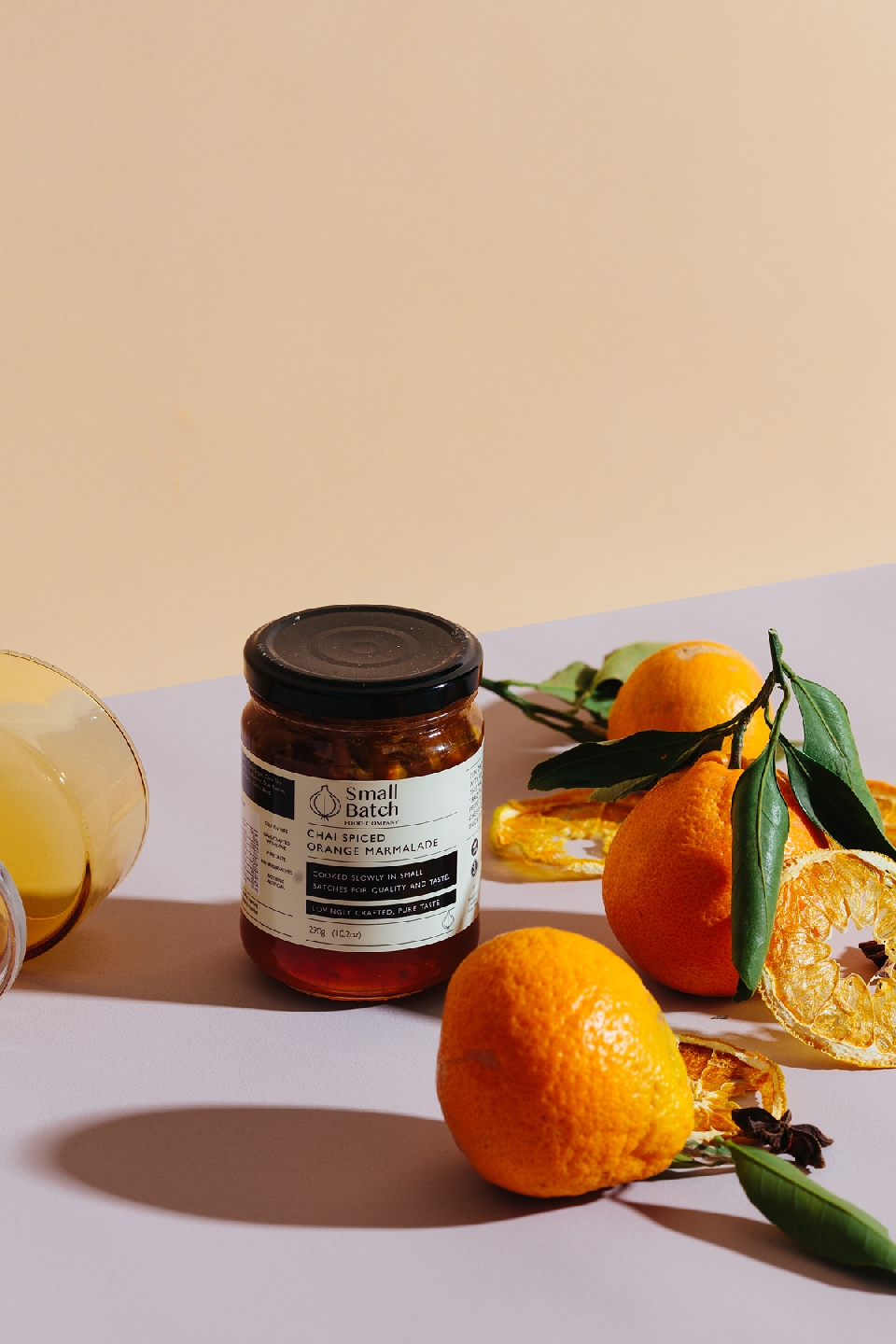 Still life of a jar of marmalade with fresh oranges and dried orange slices on a light purple and cream colored surface and background.