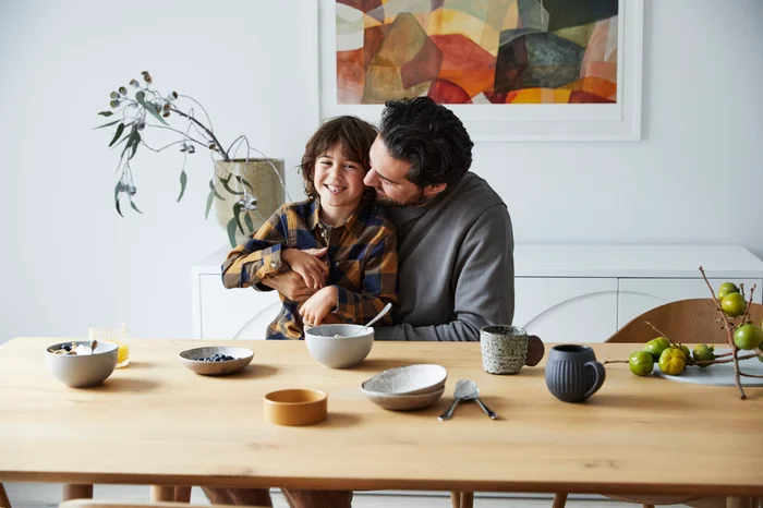 Father and child sitting together at a wooden dining table styled with ceramic bowls and fresh fruit in a bright interior space.
