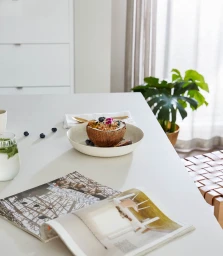 A coconut bowl filled with granola and blueberries rests on a white kitchen counter next to a magazine