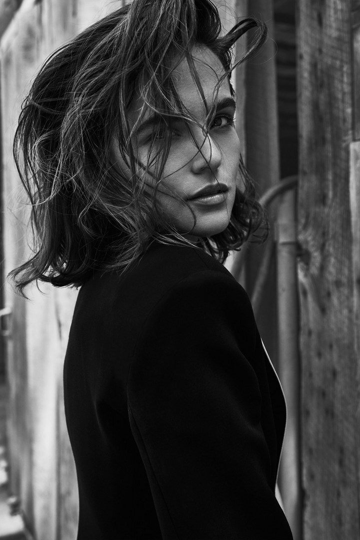Monochrome portrait of a stylish adult woman with short, wet hair wearing a dark blazer, standing outdoors against a weathered wood backdrop.