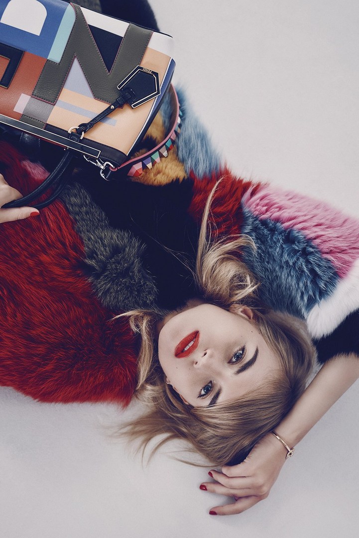 A young blonde woman lies down on a light surface wearing a multicolor fur coat and holding a geometric patterned handbag.