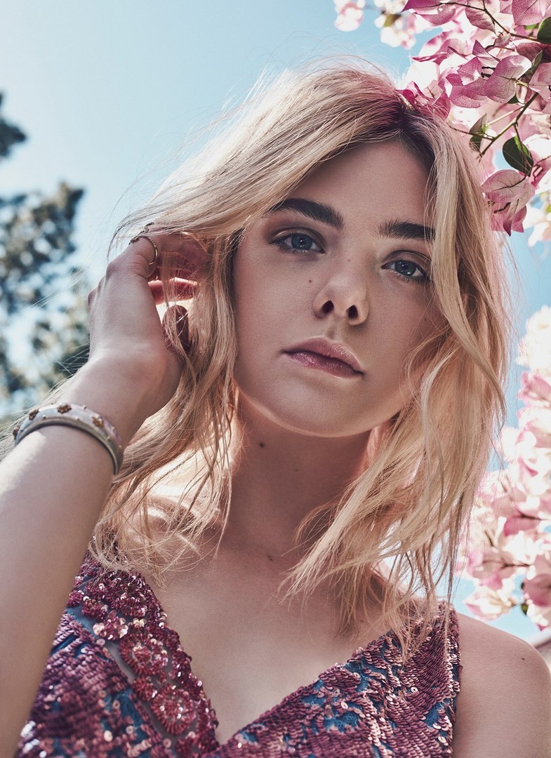 Portrait of a blonde woman with fair skin and a pink sequin top, posing outdoors under a pink-flowered tree against a light blue sky.