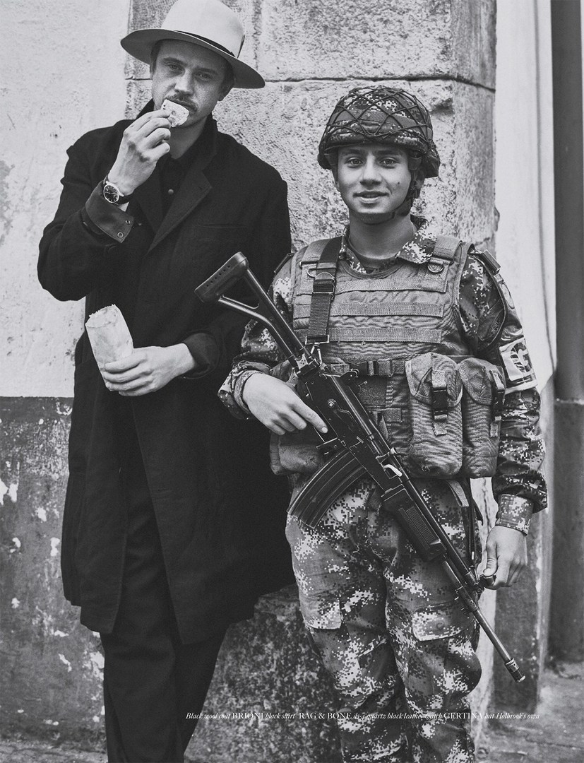 Monochrome image featuring a man in a hat eating and a soldier in camouflage holding a rifle against an old stone wall. 