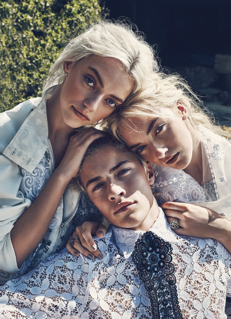 Three fair-skinned young adults pose closely together outdoors wearing matching light-blue patterned lace tops and dresses in daylight.