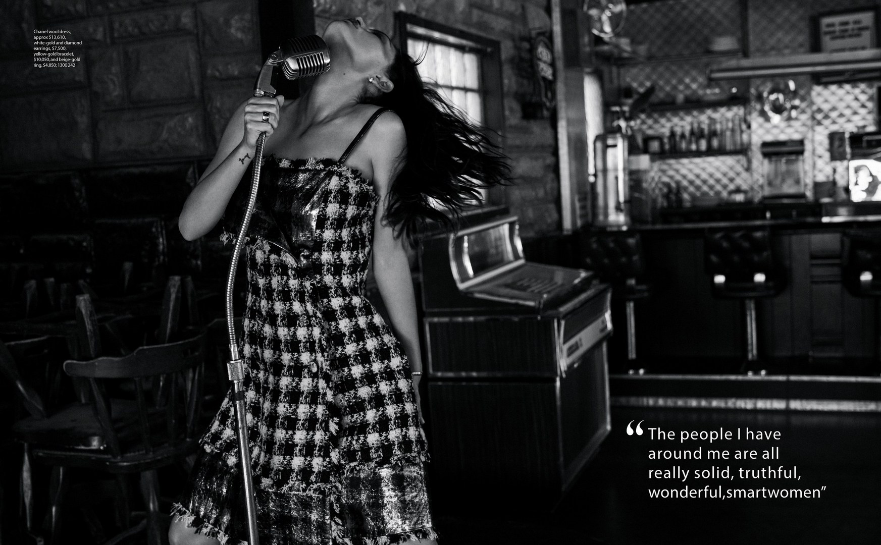An adult woman sings into a vintage microphone in black and white; she wears a patterned dress in front of a bar-like setup.