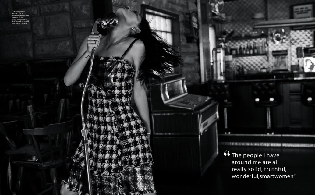 An adult woman sings into a vintage microphone in black and white; she wears a patterned dress in front of a bar-like setup.