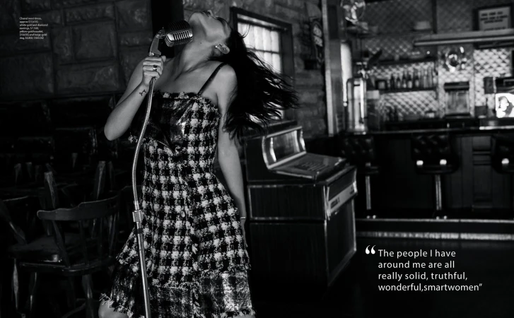 An adult woman sings into a vintage microphone in black and white; she wears a patterned dress in front of a bar-like setup.