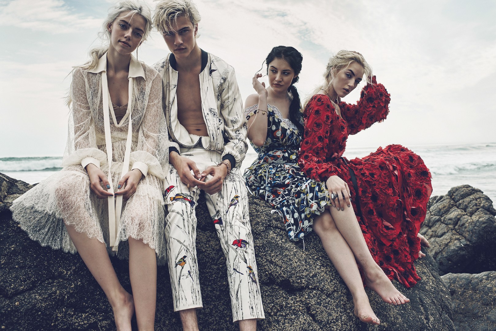 Four young adults in high-fashion, posed on a rocky beach with ocean backdrop. Focus on detailed attire and natural light.