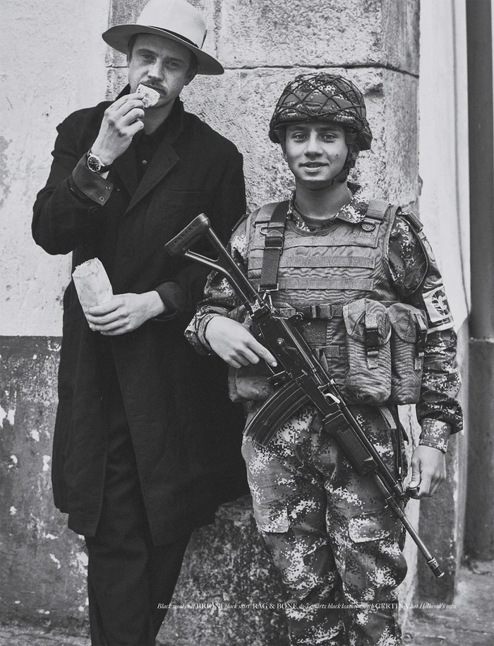 Monochrome portrait of a man in a hat eating alongside a soldier in camouflage gear holding a rifle, photographed by Beau Grealy.