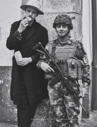 Monochrome portrait of a man in a hat eating alongside a soldier in camouflage gear holding a rifle, photographed by Beau Grealy.