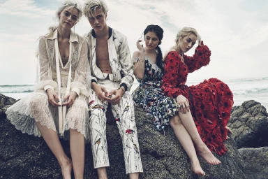 Four models seated on coastal rocks by the ocean wearing patterned clothing and sheer white lace dresses by photographer Beau Grealy