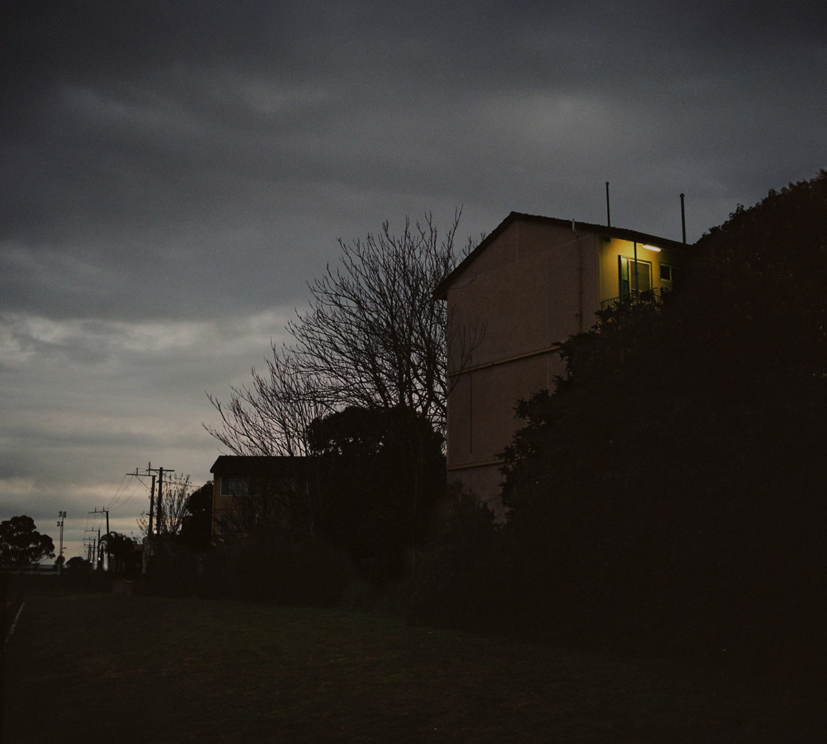 Dark twilight landscape with silhouetted apartment buildings under a heavy grey sky and a single illuminated window.
