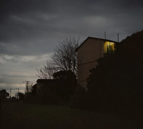 Dark twilight landscape with silhouetted apartment buildings under a heavy grey sky and a single illuminated window.