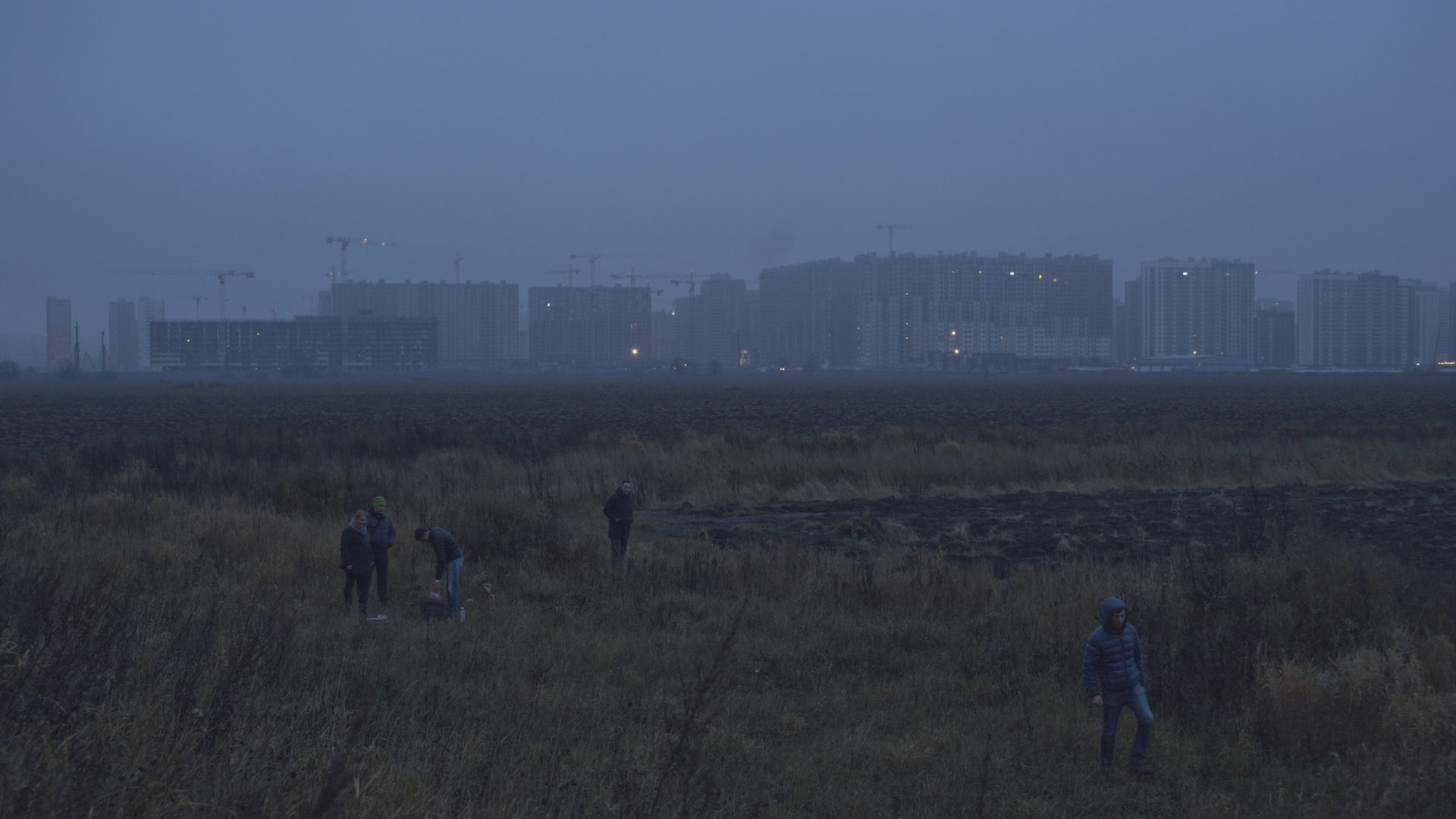 A group of people gather in a field at dusk with a distant city skyline and construction cranes visible against a hazy blue sky.