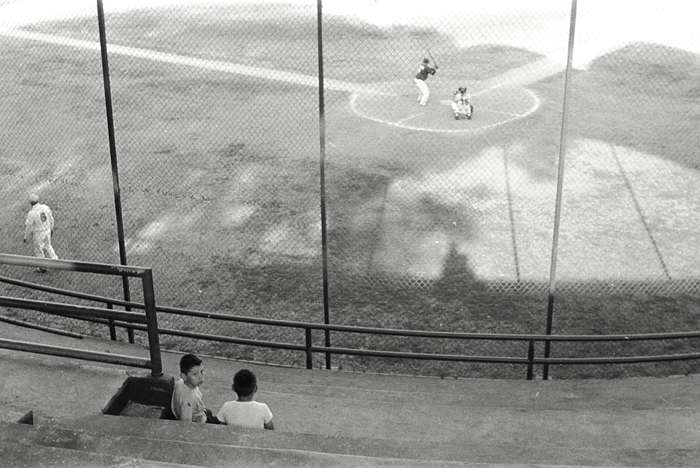 Black and white photo of children watching a baseball game from stadium seats, with baseball players and a field visible in the background.