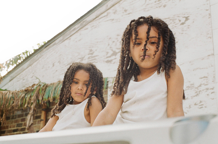 Two young girls with dark hair, wearing white tops, sit close together against a rustic building with straw details, looking serious.