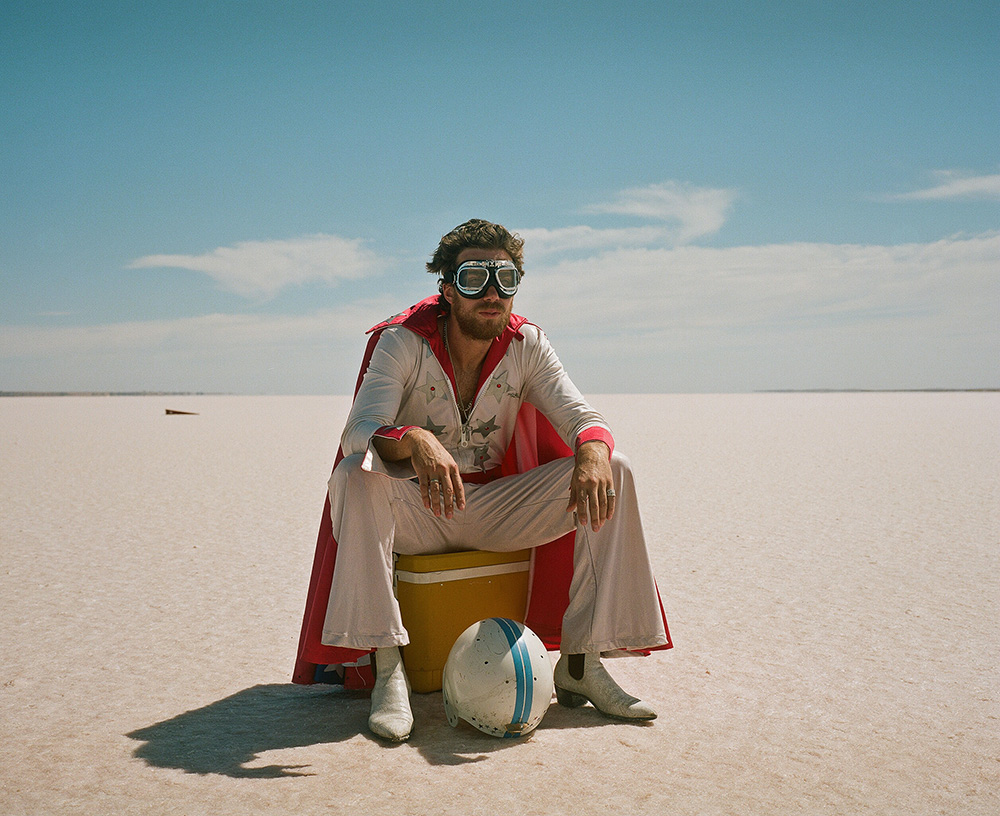 Man in white jumpsuit and cape sits on cooler on salt flats with helmet, sunglasses, and boots under clear blue sky.