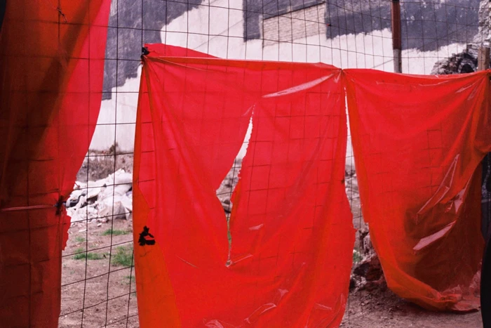 Red plastic sheets hang on a chain-link fence, partially obscuring a building under construction in an urban environment during the daytime.