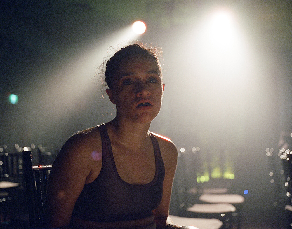 Close up of an adult woman in a tank top seated in a chair under bright stage lights with rows of empty chairs in the background.