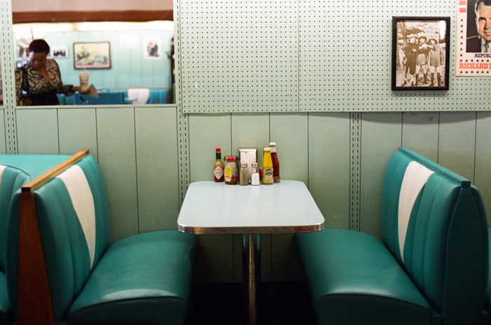 Diner booth with table, condiments, and pictures on walls. Mirror reflects a woman at counter. Cool, vintage atmosphere.